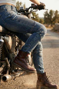 Person wearing blue jeans and brown boots sitting on a motorcycle on a road.