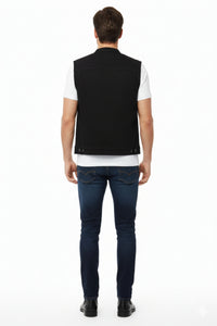 Man wearing a black vest over a white shirt with dark jeans on a white background
