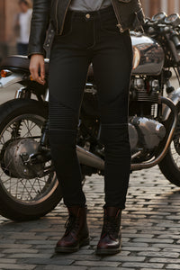 Person wearing black leather jacket, jeans, and boots standing next to a motorcycle on a cobblestone street.